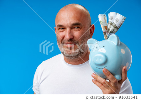 Middle-aged man holding piggy bank against blue background Middle-aged man holding piggy bank against blue background 91033541