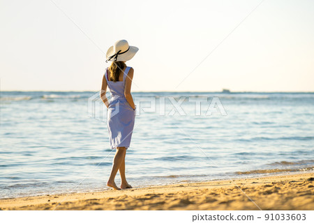 Young woman in straw hat and a dress walking alone on empty sand beach at sea shore. Lonely tourist girl looking at horizon over calm ocean surface on vacation trip. Young woman in straw hat and a dress walking alone on empty sand beach at sea shore. Lonely tourist girl looking at horizon over calm ocean surface on vacation trip. 91033603