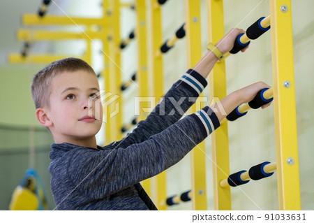 Young child boy exercising on a wall ladder bar inside sports gym room in a school. Young child boy exercising on a wall ladder bar inside sports gym room in a school. 91033631