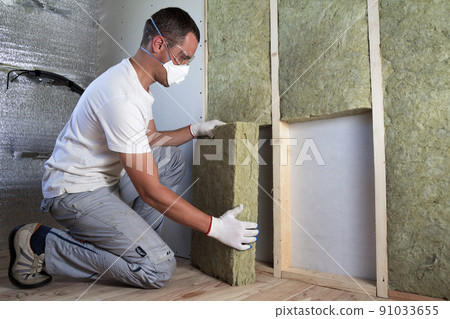 Worker in protective goggles and respirator insulating rock wool insulation in wooden frame for future house walls for cold barrier. Comfortable warm home, economy, construction and renovation concept 91033655