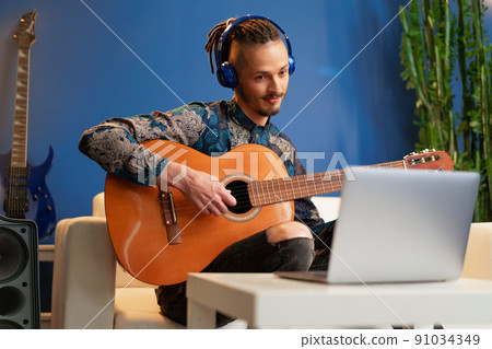 Young man sitting in his room and watching guitar tutorial on laptop Young man sitting in his room and watching guitar tutorial on laptop 91034349