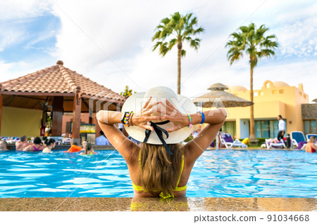 Back view of young woman with long hair wearing straw hat relaxing in summer in hotel swimming pool with blue water on a sunny day. Back view of young woman with long hair wearing straw hat relaxing in summer in hotel swimming pool with blue water on a sunny day. 91034668