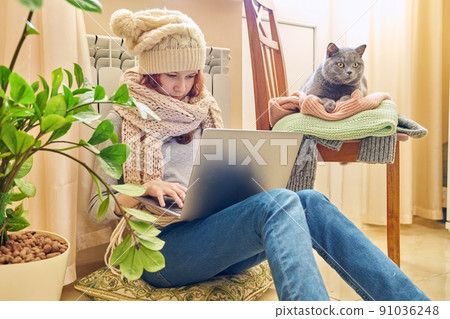 Preteen girl in scarf hat using laptop, warming near heating radiator with pet gray cat 91036248
