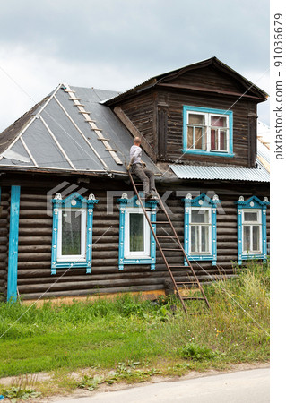 Middle-aged man sitting back on metal ladder repairing roof of old wooden brown house carved windows Middle-aged man sitting back on metal ladder repairing roof of old wooden brown house carved windows 91036679