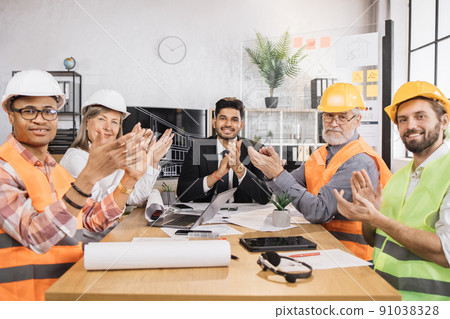 Construction team in suits and helmets sitting at table with blueprints and gadgets. 91038328