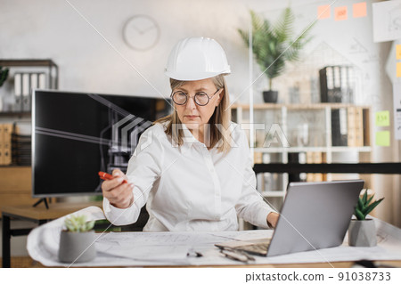 Mature architect woman or female builder sitting on the desk at workplace in modern office. Mature architect woman or female builder sitting on the desk at workplace in modern office. 91038733