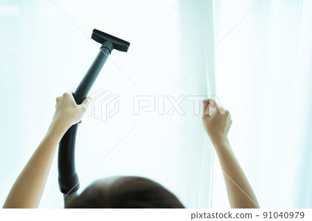 Asian young woman using vacuum machine to cleanup the dust on curtain in living room close up. Housework and chores in daily life concept. Housekeeper cleaning a white curtain. 91040979