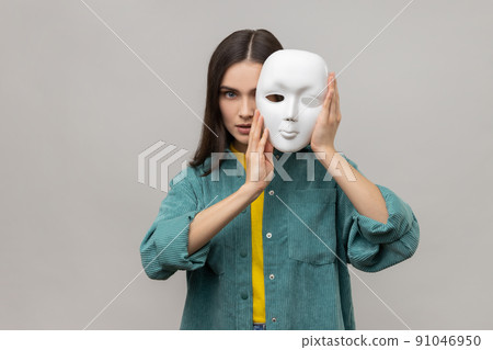 Portrait of strict bossy woman covering half of face with white mask, multiple personality disorder, wearing casual style jacket. Indoor studio shot isolated on gray background. 91046950