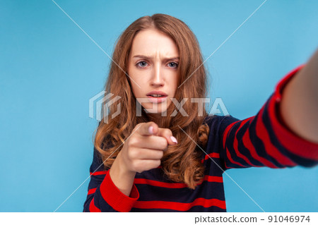 Serious woman wearing striped casual style sweater, taking selfie POV, looking at camera with srtict expression and pointing finger at you. Indoor studio shot isolated on blue background. 91046974