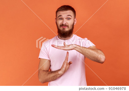 Portrait of bearded man with frowning face showing timeout gesture, tired of lot of homework, extra classes, wearing pink T-shirt. Indoor studio shot isolated on orange background. 91046978