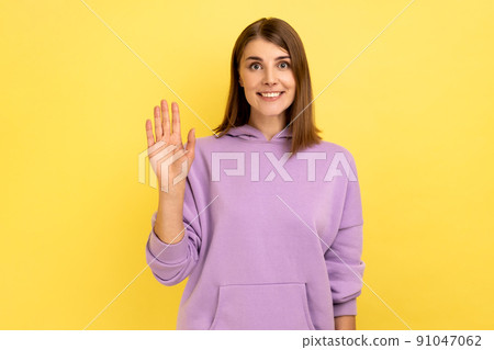 Portrait of friendly positive young woman smiles toothily, raises palm greets friend, saying hello or goodbye, wearing purple hoodie. Indoor studio shot isolated on yellow background. 91047062