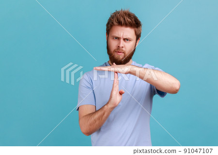 I need more time. Portrait of bearded man showing time out gesture, looking with imploring eyes, hurry to meet deadline, begging for more time. Indoor studio shot isolated on blue background. I need more time. Portrait of bearded man showing time out gesture, looking with imploring eyes, hurry to meet deadline, begging for more time. Indoor studio shot isolated on blue background. 91047107