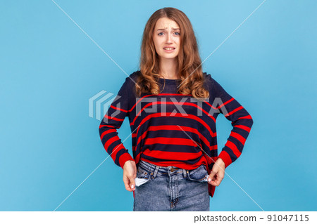 I'm bankrupt. Unhappy poor woman, turning empty pockets inside out and looking frustrated by overspend, lack of money, wearing striped casual sweater. Indoor studio shot isolated on blue background. I'm bankrupt. Unhappy poor woman, turning empty pockets inside out and looking frustrated by overspend, lack of money, wearing striped casual sweater. Indoor studio shot isolated on blue background. 91047115