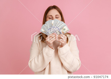 Portrait of rich greedy blond woman holding big fan of dollars bills and smelling money with satisfied expression, wearing white sweater. Indoor studio shot isolated on pink background. Portrait of rich greedy blond woman holding big fan of dollars bills and smelling money with satisfied expression, wearing white sweater. Indoor studio shot isolated on pink background. 91047174