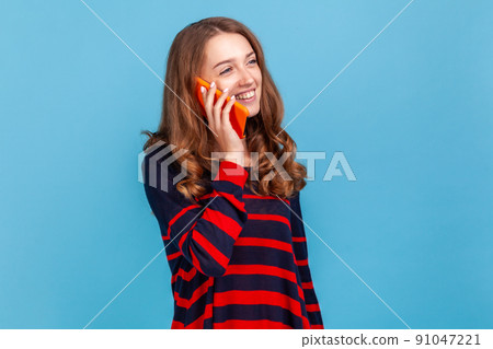 Portrait of positive happy woman wearing striped casual style sweater, having pleasant conversation, standing and talking phone with smile.Indoor studio shot isolated on blue background. Portrait of positive happy woman wearing striped casual style sweater, having pleasant conversation, standing and talking phone with smile.Indoor studio shot isolated on blue background. 91047221