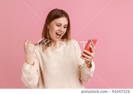 Extremely happy excited blond woman standing with cell phone in hands and clenching fist, celebrating her victory, great news, wearing white sweater. Indoor studio shot isolated on pink background. Extremely happy excited blond woman standing with cell phone in hands and clenching fist, celebrating her victory, great news, wearing white sweater. Indoor studio shot isolated on pink background. 91047228