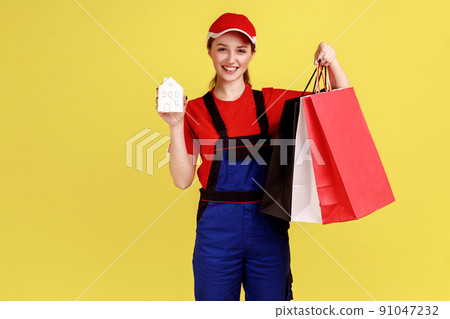 Portrait of courier woman standing with shopping bags and paper house in hands, safe door-to-door delivery, wearing overalls and red cap. Indoor studio shot isolated on yellow background. Portrait of courier woman standing with shopping bags and paper house in hands, safe door-to-door delivery, wearing overalls and red cap. Indoor studio shot isolated on yellow background. 91047232
