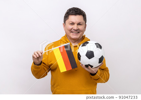Middle aged man with toothy smile holding flag of germany and soccer black and white classic ball and watching match, wearing urban style hoodie. Indoor studio shot isolated on white background. Middle aged man with toothy smile holding flag of germany and soccer black and white classic ball and watching match, wearing urban style hoodie. Indoor studio shot isolated on white background. 91047233