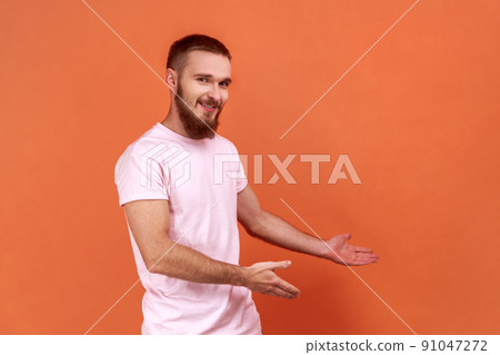Portrait of kind-hearted generous bearded man showing welcome gesture and empty space on wall for your best advertising, wearing pink T-shirt. Indoor studio shot isolated on orange background. 91047272