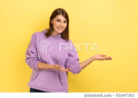 Please, take for free. Generous friendly pretty woman welcoming with wide open arms and smiling kindly, happy to embrace you, wearing purple hoodie. Indoor studio shot isolated on yellow background. 91047301