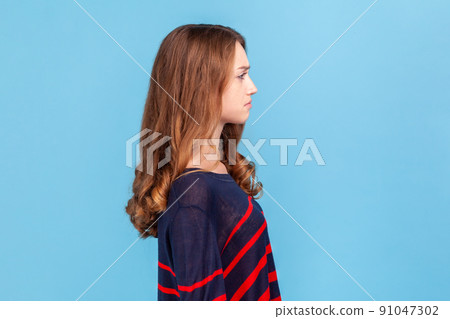 Side view of sad upset young woman wearing striped casual style sweater standing looking ahead, being in bad mood, suffering depression. Indoor studio shot isolated on blue background. 91047302