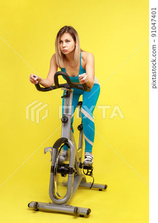 Full length of beautiful blonde woman doing cardio workout on exercise bike, loosing weigh or improving her body shape, wearing blue sportswear. Indoor studio shot isolated on yellow background. 91047401