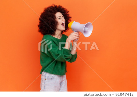 Side view portrait of woman with Afro hairstyle wearing green casual style sweater protesting or making announcement, screaming in megaphone. Indoor studio shot isolated on orange background. Side view portrait of woman with Afro hairstyle wearing green casual style sweater protesting or making announcement, screaming in megaphone. Indoor studio shot isolated on orange background. 91047412
