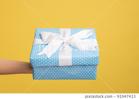 Closeup side view of woman hand holding out blue gift box with ribbon, giving present on holiday, bonuses and surprises concept. Indoor studio shot isolated on yellow background. 91047413