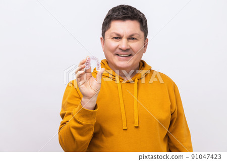 Portrait of smiling middle aged man with dark hair holding transparent plastic braces, mouth guard, wearing urban style hoodie. Indoor studio shot isolated on white background. Portrait of smiling middle aged man with dark hair holding transparent plastic braces, mouth guard, wearing urban style hoodie. Indoor studio shot isolated on white background. 91047423