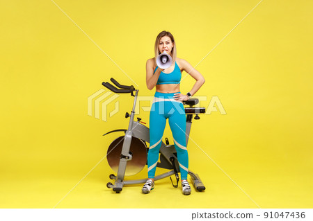 Full length portrait of sporty woman holding megaphone and making announce, inviting potential clients to fitness club, wearing blue sportswear. Indoor studio shot isolated on yellow background. Full length portrait of sporty woman holding megaphone and making announce, inviting potential clients to fitness club, wearing blue sportswear. Indoor studio shot isolated on yellow background. 91047436