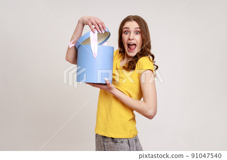 Amazed teenager girl in yellow casual T-shirt opening gift box, having excited surprised expression, unpacking birthday present surprise. Indoor studio shot isolated on gray background. 91047540
