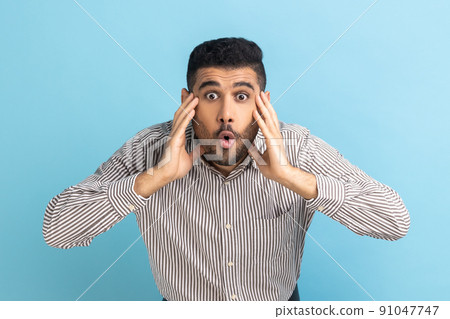 Wow, it's unbelievable. Surprised businessman staring at camera with widely open mouth and big eyes, raising arms in amazement, wearing striped shirt. Indoor studio shot isolated on blue background. 91047747