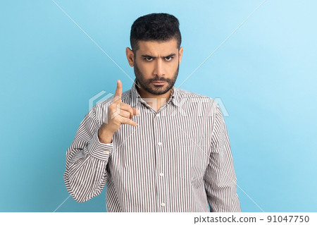 Portrait of serious strict bearded businessman standing with admonishing gesture, scolding for mistake and warning, wearing striped shirt. Indoor studio shot isolated on blue background. Portrait of serious strict bearded businessman standing with admonishing gesture, scolding for mistake and warning, wearing striped shirt. Indoor studio shot isolated on blue background. 91047750