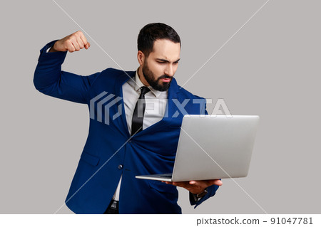 Man punching laptop screen, looking with furious mad expression, boxing threatening to hit while having online conversation, wearing official style suit. Indoor studio shot isolated on gray background 91047781