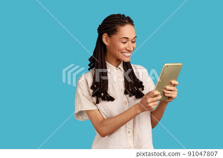 Creative smiling woman with black dreadlocks works on future project, using tablet for checking social networks, wearing white shirt. Indoor studio shot isolated on blue background. Creative smiling woman with black dreadlocks works on future project, using tablet for checking social networks, wearing white shirt. Indoor studio shot isolated on blue background. 91047807