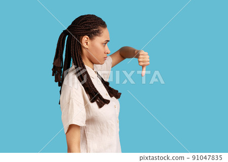 Side view of discontent woman with black dreadlocks showing disapproval sign, keeps thumb down, expresses dislike, frowns face in discontent. Indoor studio shot isolated on blue background. Side view of discontent woman with black dreadlocks showing disapproval sign, keeps thumb down, expresses dislike, frowns face in discontent. Indoor studio shot isolated on blue background. 91047835