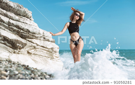 a girl in a swimsuit poses in the spray of waves at a white rock on the seashore a girl in a swimsuit poses in the spray of waves at a white rock on the seashore 91050281