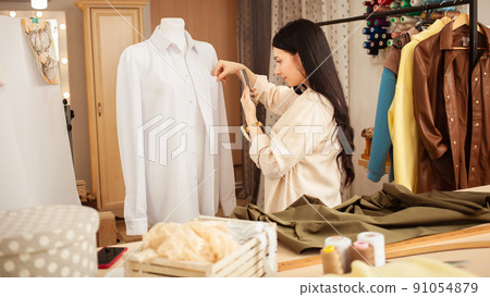 Seamstress in sewing studio, indoors. Beautiful woman, cozy interior. young entrepreneur. photographs finished product 91054879