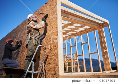 Carpenters hammering nail into OSB panel on the wall of future cottage. Men workers building wooden frame house in the Scandinavian style barnhouse. Carpentry and construction concept. 91057103