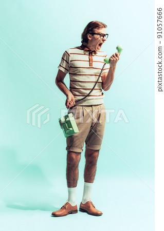 Portrait of young emotive man in retro style outfit posing with old-fashioned phone isolated over light blue studio background. Screaming 91057666