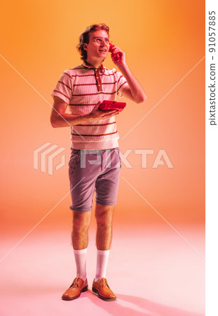 Full-length portrait of stylish young man in retro outfit posing with vintage phone isolated over orange background in neon light Full-length portrait of stylish young man in retro outfit posing with vintage phone isolated over orange background in neon light 91057885