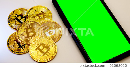 Bitcoin coins in golden metal with a smarthphone beside it, green screen area copy space 91068020