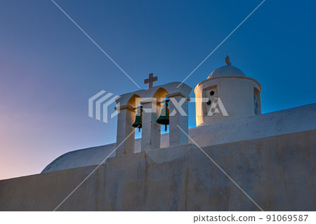 Whitewashed Greek Orthodox chapel, small belfry with cross and bell, roof and dome at sunset sky background. Close-up shot. Milos island, Greece 91069587
