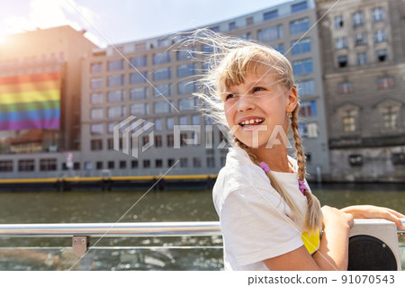 Portrait of cute adorable blond child girl stand on board river Spree Cruise trip boat with LGBTQ flag poster on building on background on brigh sunny day Berlin . Pride day fest celebraion Germany 91070543