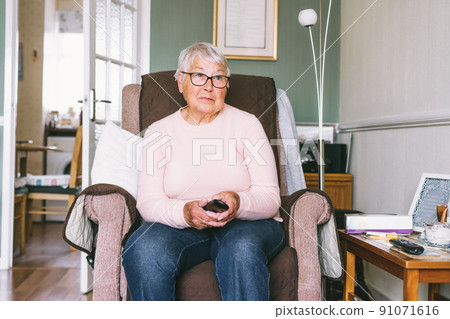 Portrait of retired elderly woman in comfortable armchair with TV remote control and watching television channel in living room. Retirement concept. Selective focus. Portrait of retired elderly woman in comfortable armchair with TV remote control and watching television channel in living room. Retirement concept. Selective focus. 91071616