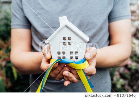 Close up male hands holding house statuette in hands with blue and yellow ribbons, the colors of the Ukrainian flag. Home for refugees from Ukraine, help for families. Selective focus, copy space. Close up male hands holding house statuette in hands with blue and yellow ribbons, the colors of the Ukrainian flag. Home for refugees from Ukraine, help for families. Selective focus, copy space. 91071646