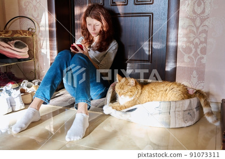 Preteen girl sitting on floor at home using smartphone, with sleeping pet cat Preteen girl sitting on floor at home using smartphone, with sleeping pet cat 91073311