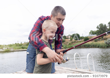 Father and son fishing. Dad shows his son how to hold the spinning and spin the reel. Fishing training on a pond or river. Caring parent concept. Father and son fishing. Dad shows his son how to hold the spinning and spin the reel. Fishing training on a pond or river. Caring parent concept. 91073979