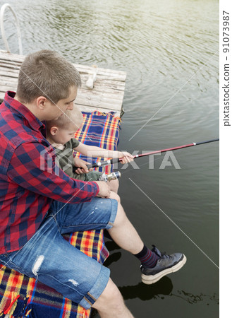 Father and son fishing. Dad shows his son how to hold the spinning and spin the reel. Fishing training on a pond or river. Caring parent concept. Father and son fishing. Dad shows his son how to hold the spinning and spin the reel. Fishing training on a pond or river. Caring parent concept. 91073987