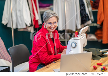 Woman smiling at laptop screen sitting at sewing machine Woman smiling at laptop screen sitting at sewing machine 91075743